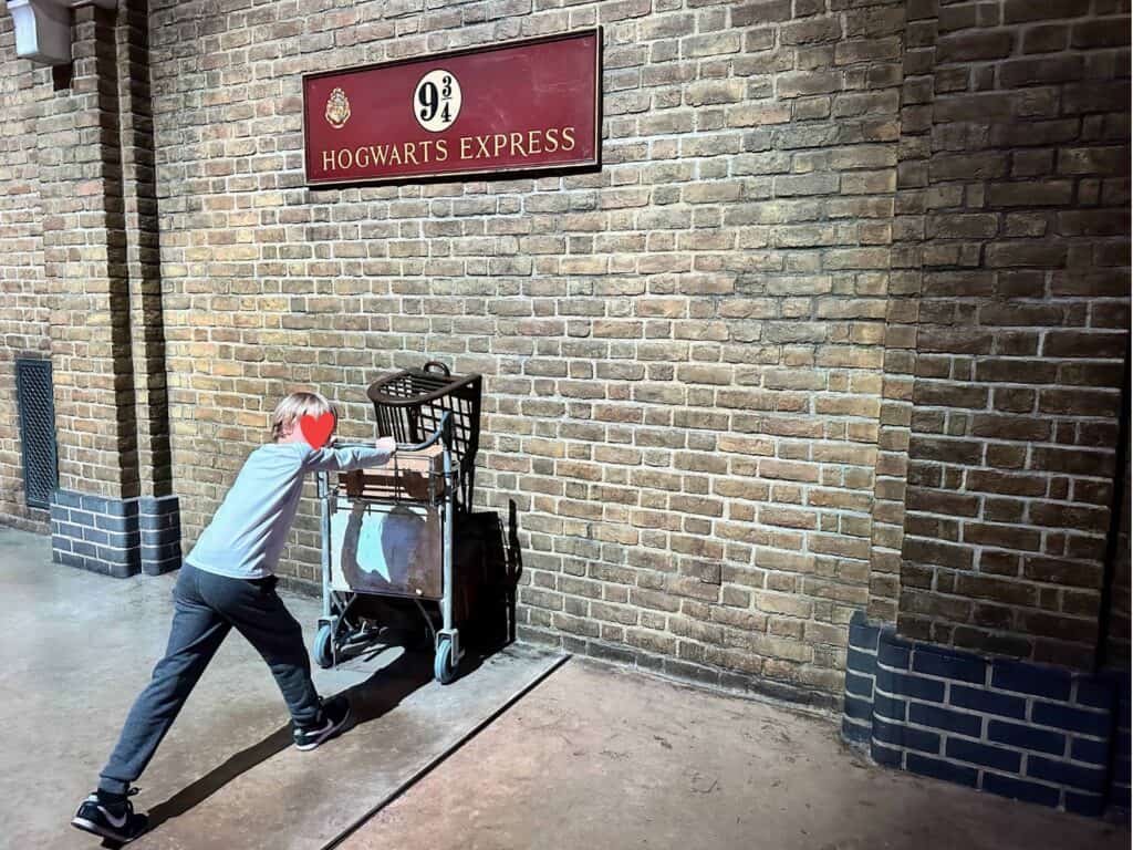 Boy pushes luggage cart into brick wall. Above is a sign for 9 ¾ Hogwarts Express.