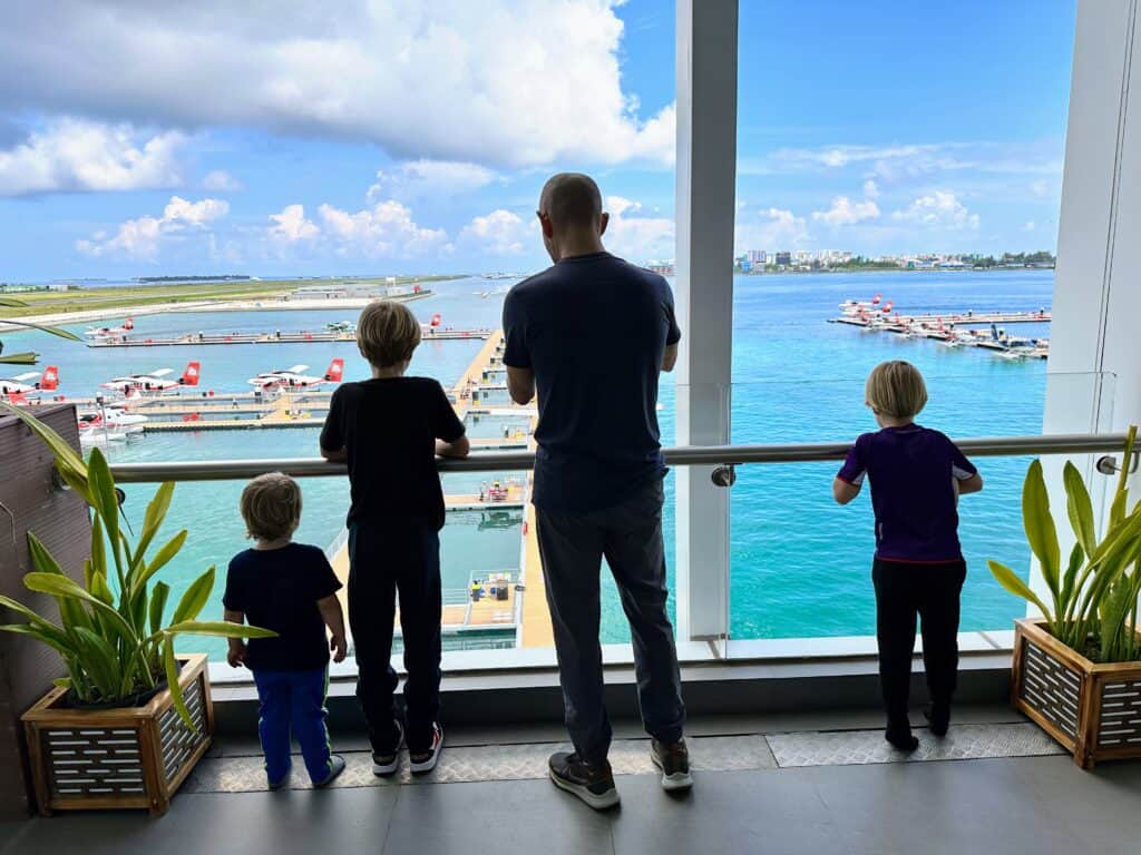 Father and sons look out floor-to-ceiling windows and watch seaplane landings and takeoffs at Malé airport in the Maldives.