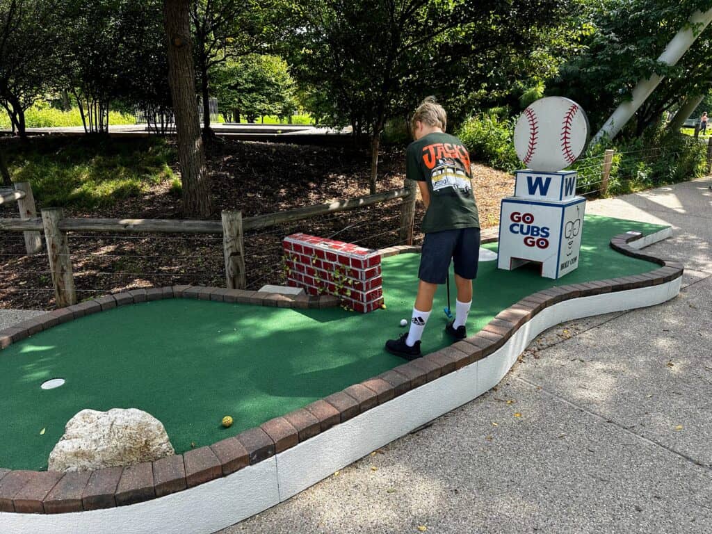 Boy putts at mini golf in Chicago next to a sign that says Go Cubs Go with a baseball.