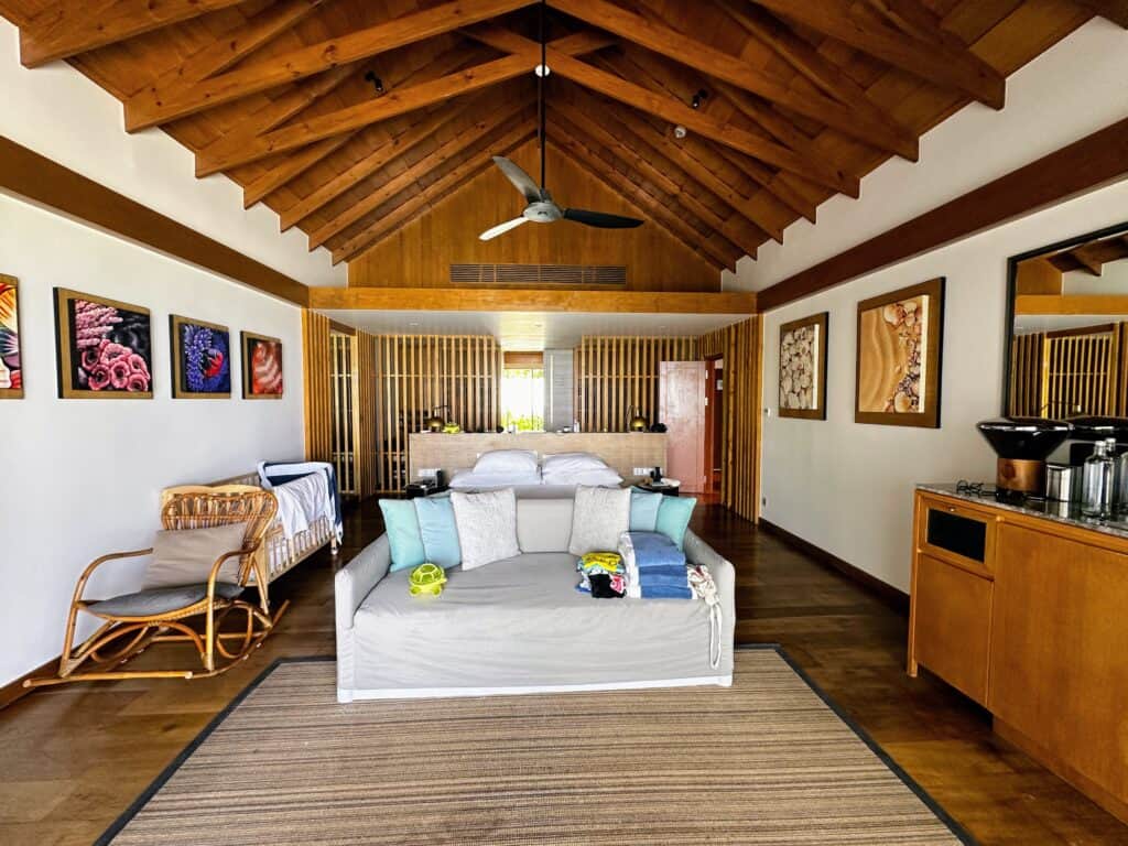 Bedroom area with bed, large couch, minibar area on right, chair and crib on left. Ceiling is wooden with a fan. Colorful marine life paintings on walls.