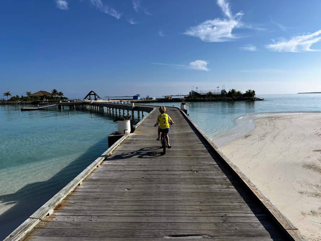 Boy in yellow shirt rides onto wooden jetty over turquoise water at Emerald Faarufushi Resort in the Maldives.