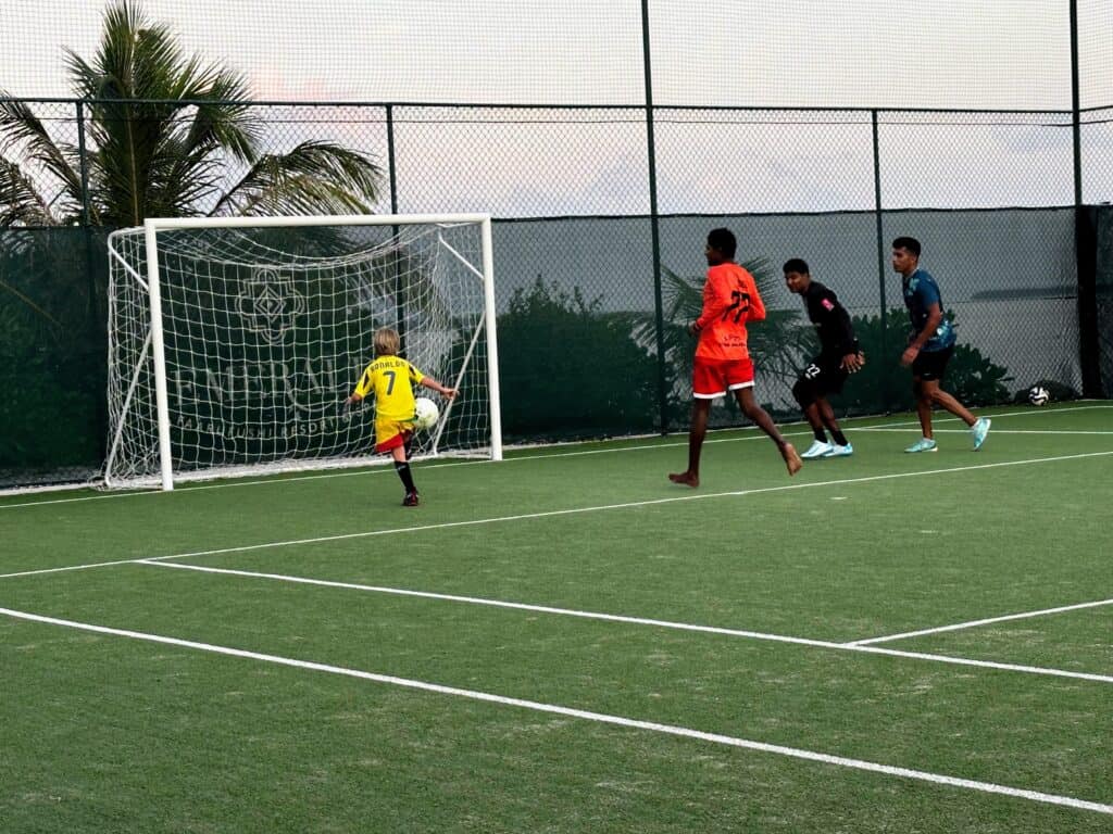 Boy in Ronaldo jersey kicks soccer ball into goal at at Emerald Faarufushi resort in the Maldives. Staff is on the right.