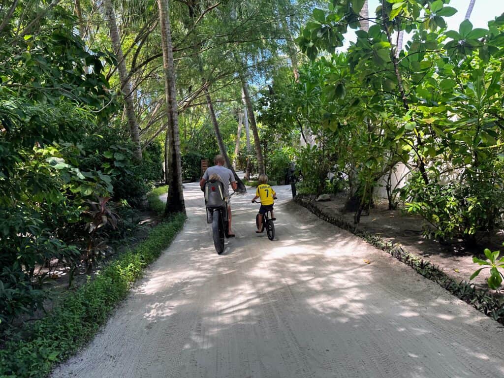 Father cycles with two sons on a dirt path at Emerald Faarufushi Resort in the Maldives. There is foliage on either side of the path.