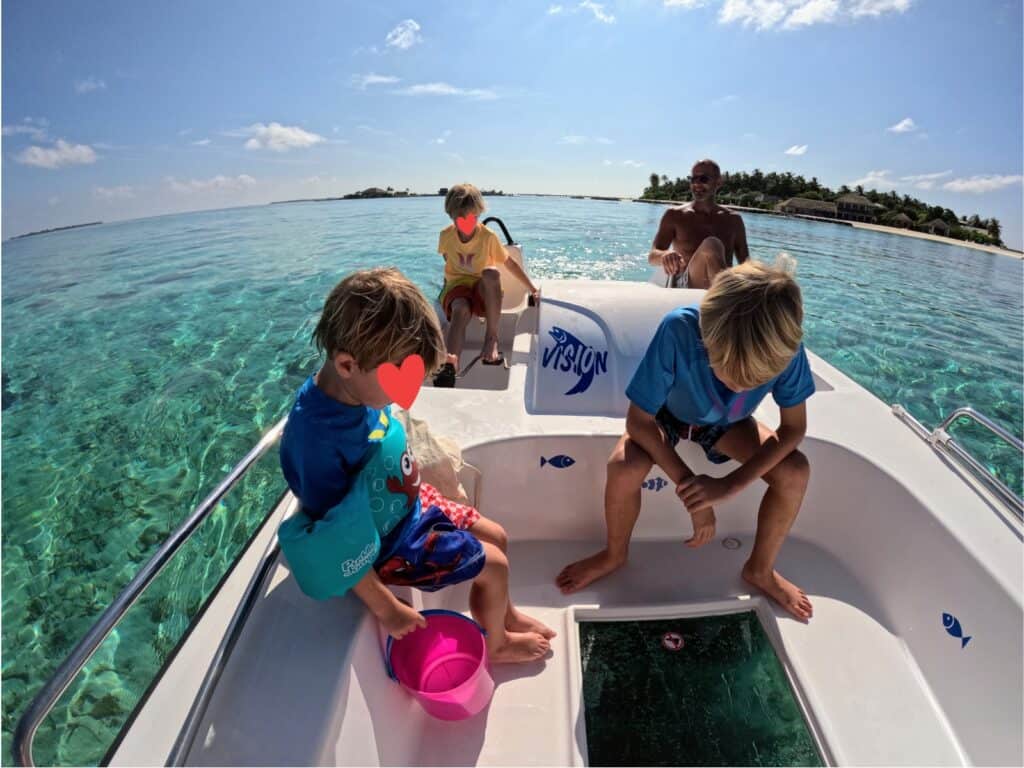 Boys look at fish through glass bottom of boat while father and other son pedal. Turquoise water and the island in the background.