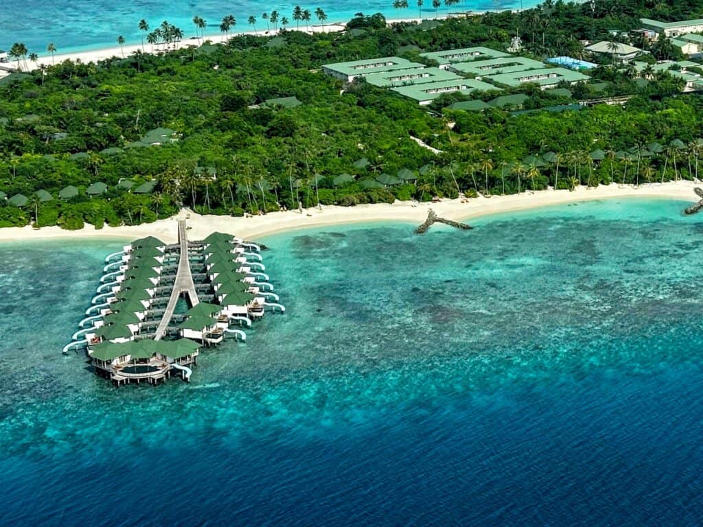 View of water villas with water slides coming off of the front part of a resort island in the Maldives. It's Siyam World. The island is lush with palm trees. There are green-roofed buildings in the center of the island.