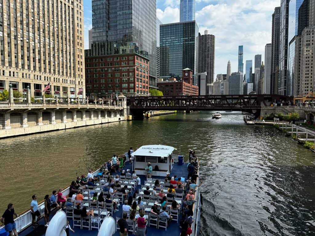 Large boat with seats and passengers listening to a guide. They are on the river in downtown Chicago. Skyscrapers on either side and up ahead.