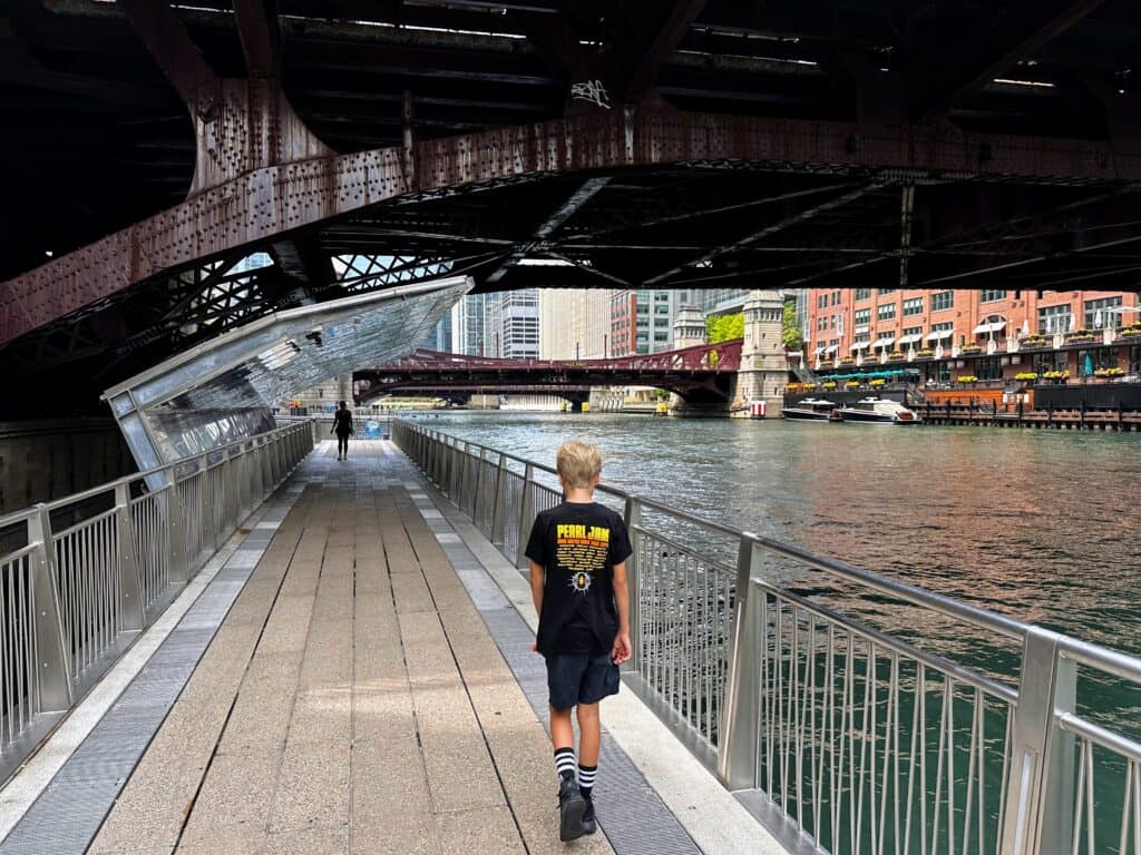 Boy wearing black Pearl Jam t-shirt walks on pedestrian path to left of river in Chicago. He's about to walk under a bridge.
