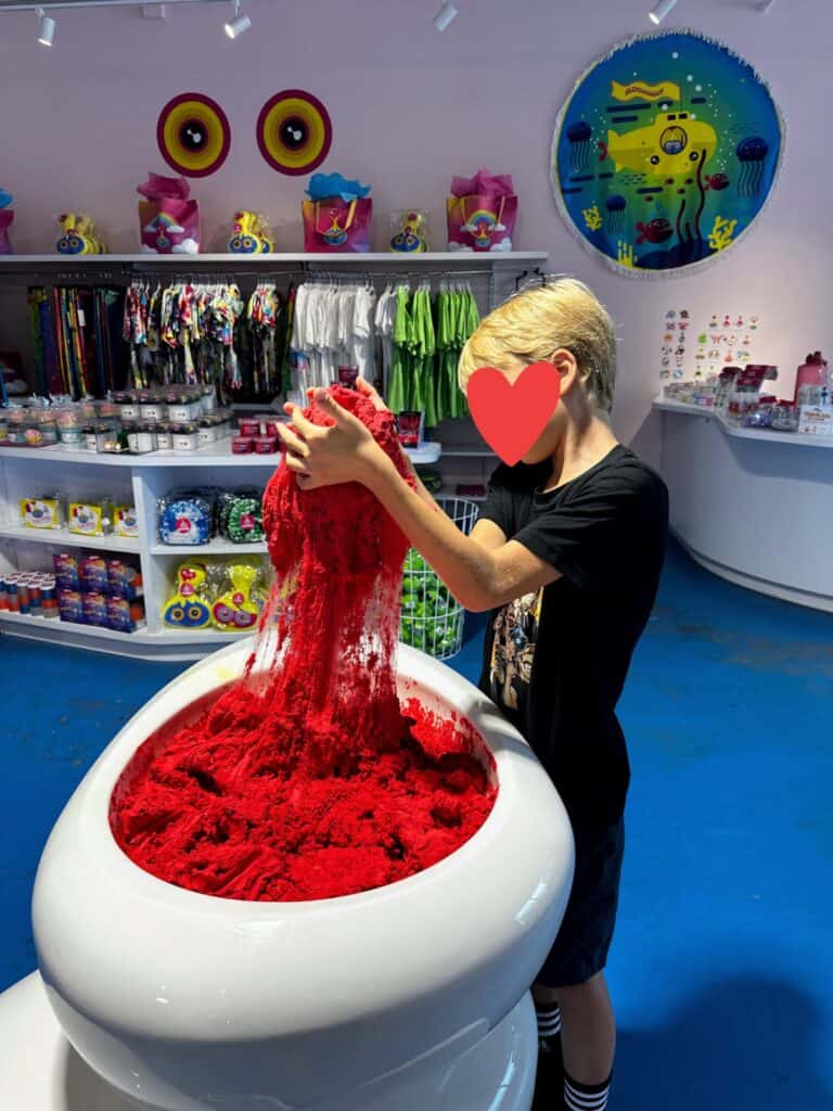 Boy stretches red slime in a white tub with gift shop shelves in background at the Sloomoo Institute in Chicago, Illinois.