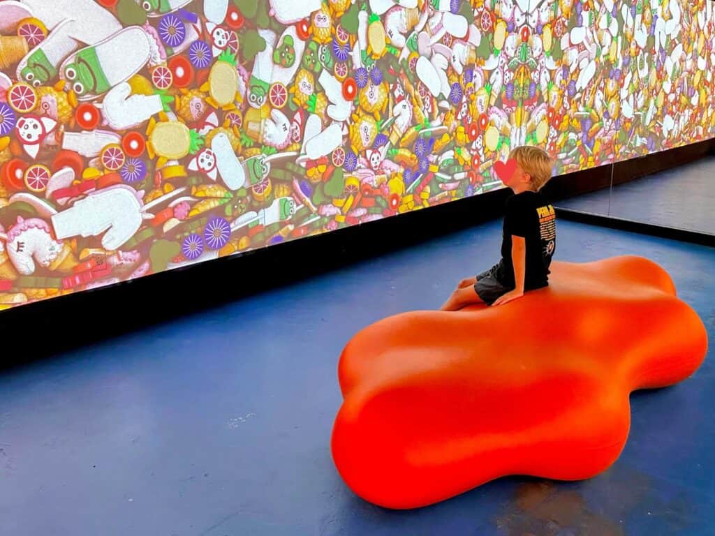 Boy sits on red blob cushion in front of screen with colorful toys on candies at the Sloomoo Institute in Chicago.