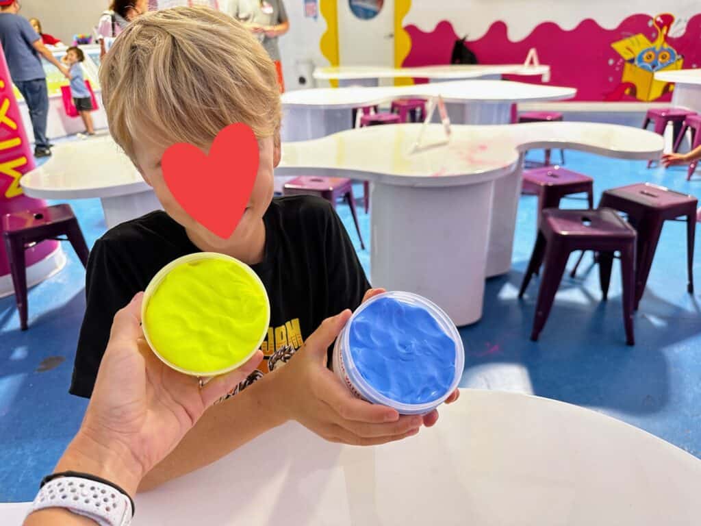 Boy holds up his blue slime and another hand holds up yellow slime at the Sloomoo Institute in Chicago, Illinois. He's sitting at a white table in a large room with other white tables and pink stools.