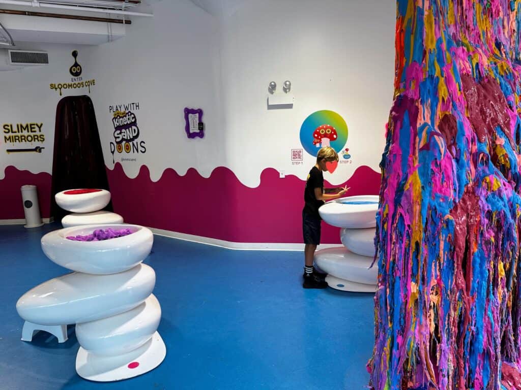 Boy plays in a bin of slime in a room with other slime bins. Slime wall with dripping, colorful slime on right.
