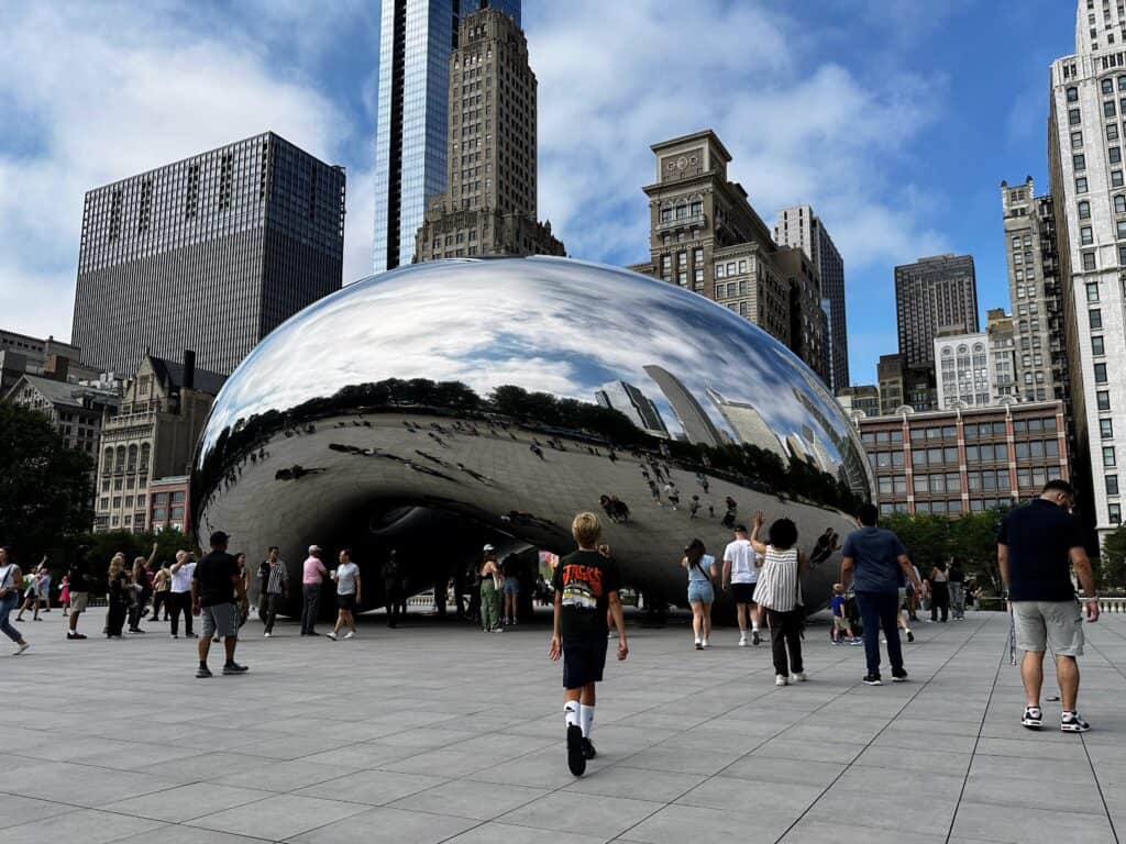 Boy walks toward the Bean in Chicago. Other people are crowded near it and there are skyscrapers behind it.