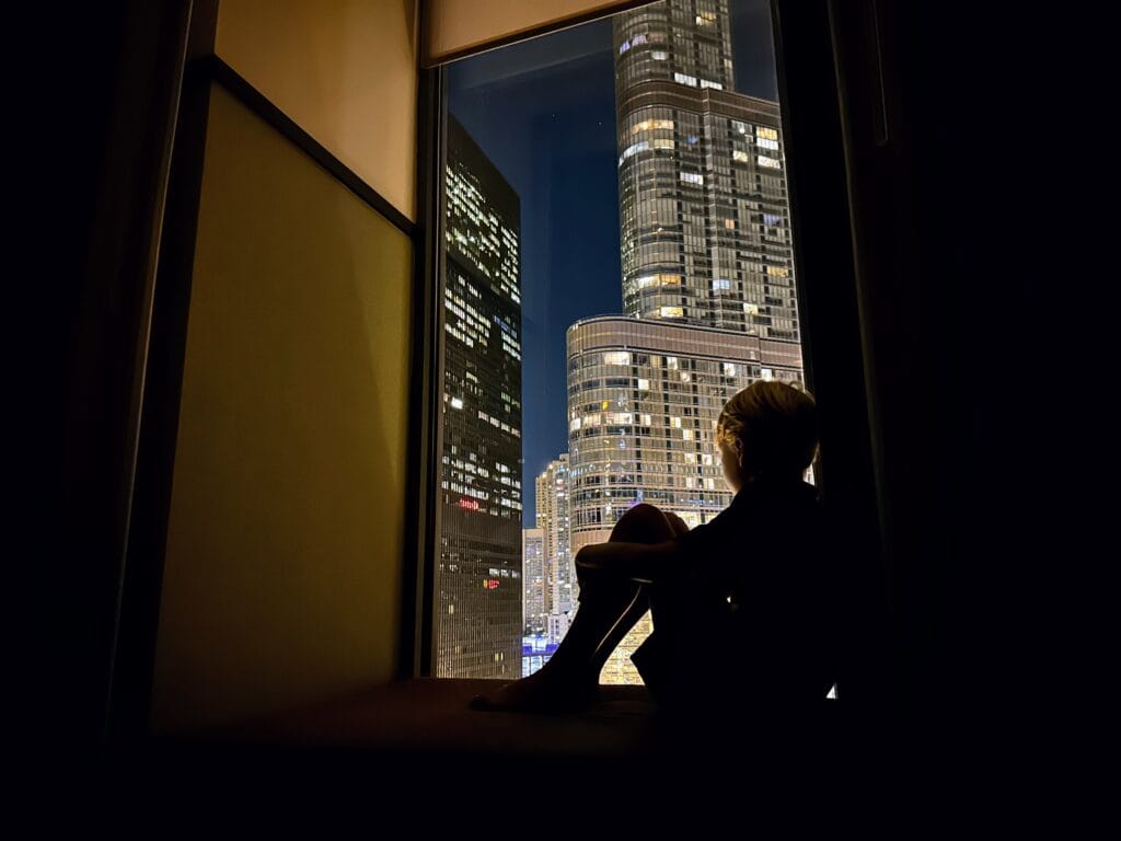 Boy sitting in a windowseat in a hotel in Chicago at night. Skyscrapers are lit up against the dark sky.