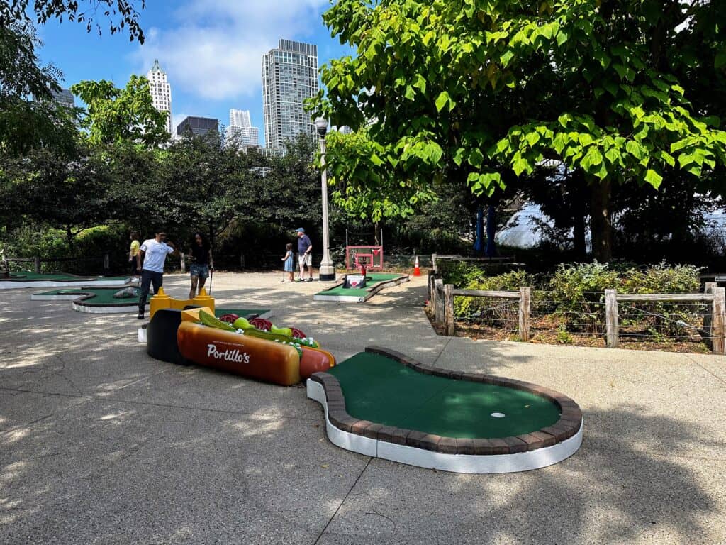 Hot dog hole at mini golf course in Chicago. Trees on either side and skyscrapers in background. A few people playing mini golf.