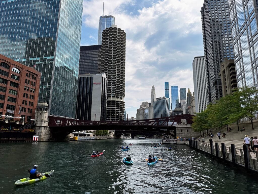 Kayakers on the river in Chicago. Skyscrapers on either side. Bridge up ahead.