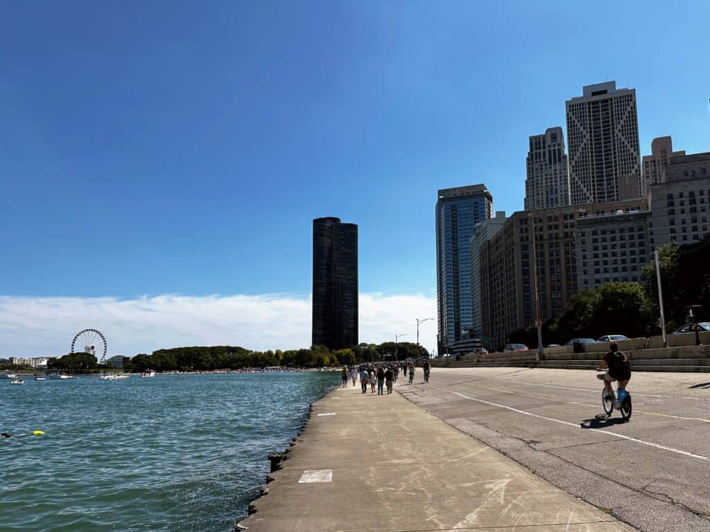 People walk and cycle along the waterfront of Lake Michigan in Chicago. Lake on left, Navy Pier with ferris wheel in back right, and tall buildings ahead and to right.