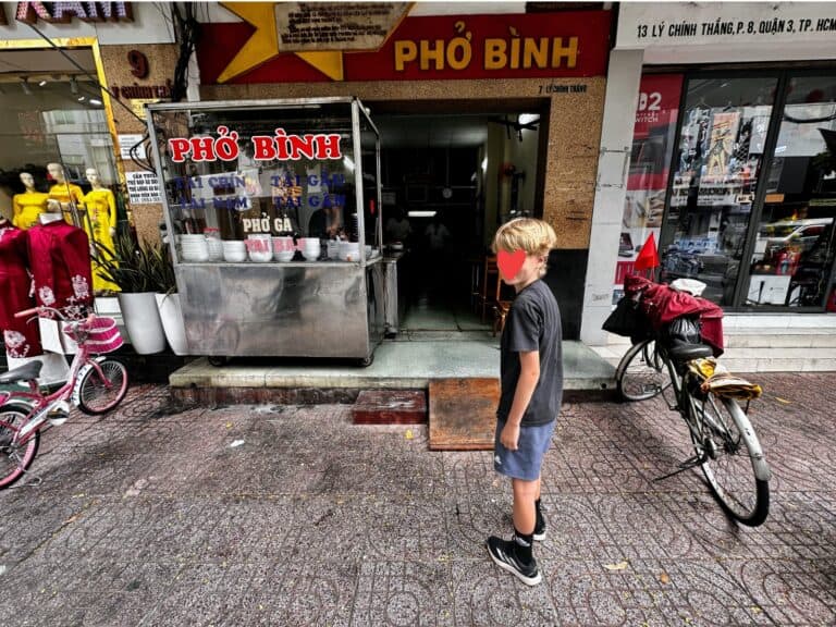 Boy walking into Pho Binh shop in Ho Chi Minh City, Vietnam. Bicycles parked out front. Window shows bowls stacked.