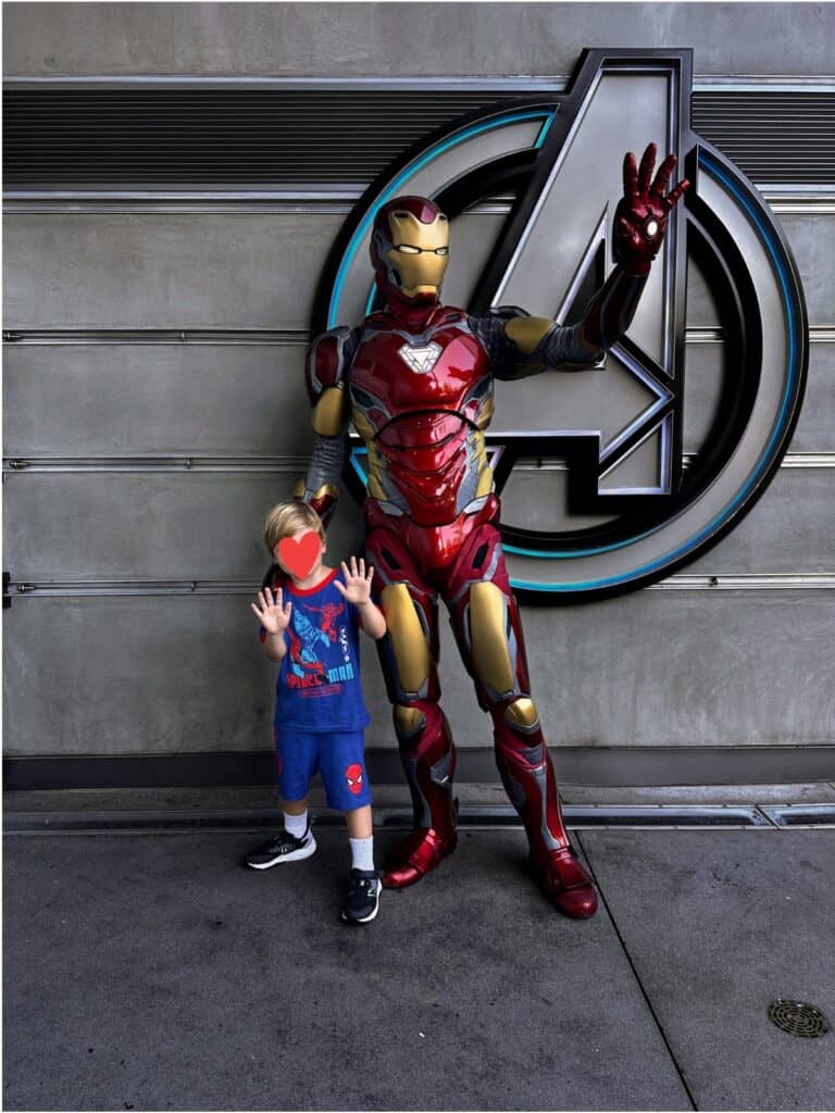 Boy poses with Iron Man at Disney California Adventure. They're standing in front of a concrete wall with a large Avengers 'A' logo.