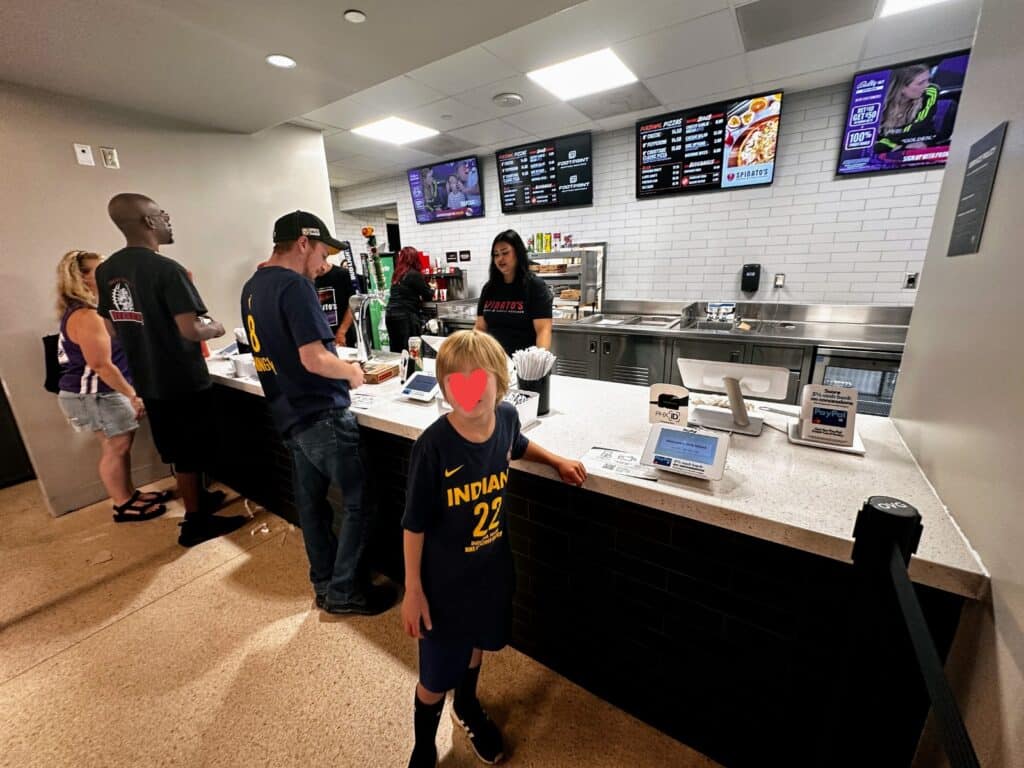 Boy stands at a concessions counter inside the basketball arena at a Phoenix Mercury WNBA game. Other people are being served by employees. Menu signs on back wall.