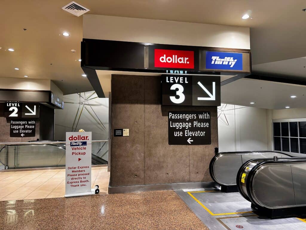Inside the car rental center at the Phoenix airport. Escalator going down on right. Signs next to escalator point toward Level 3, dollar, and Thrifty. Sign also points toward elevator to the left.