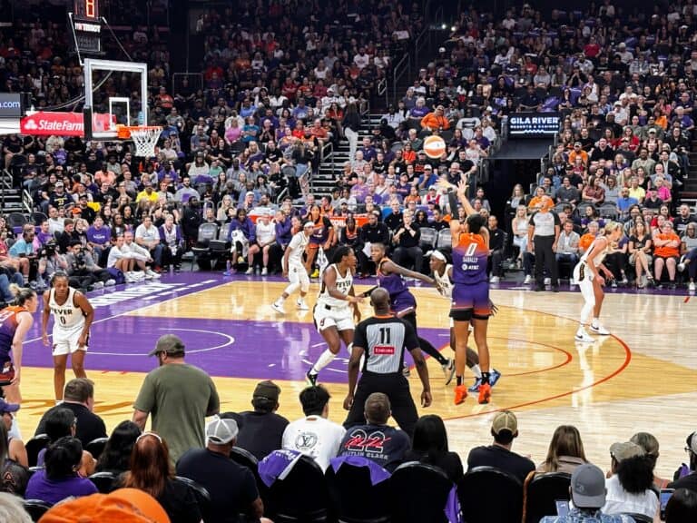 Satou Sabally shoots the ball from the 3-point line while Phoenix Mercury and Indiana Fever players look on from the court. The stands are full of fans watching the play.