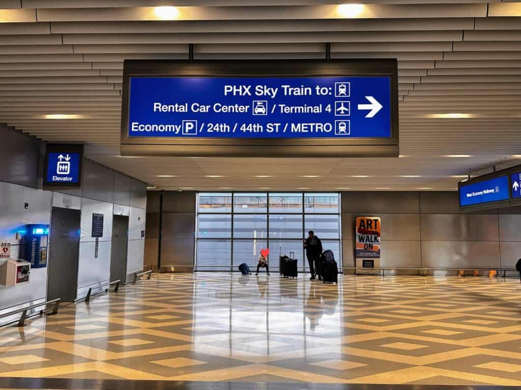 Inside terminal 3 in the Phoenix airport. Blue sign with arrow points to PHX Sky Train to Rental Car Center / Terminal 4 / Economy Parking and other public transport. Windows at end of area and two people are against the window with their luggage.