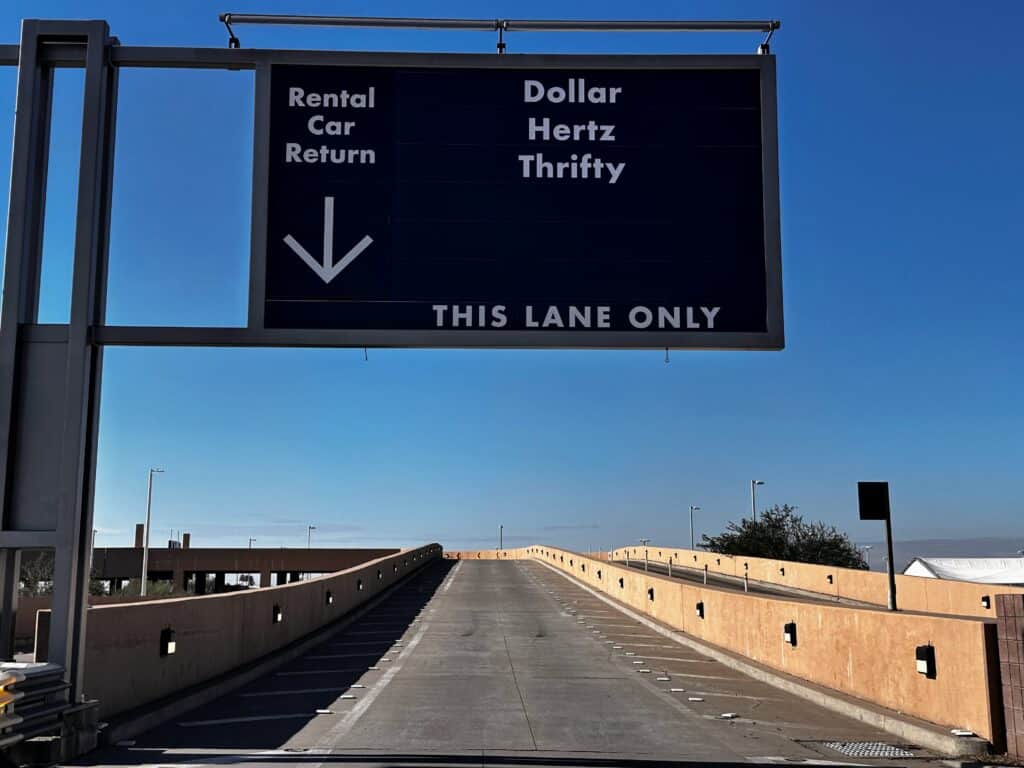 Road gently goes uphill on a ramp. Sign above road points toward 'Rental Car Return, Dollar, Hertz, Thrifty' and also says 'This Lane Only.'