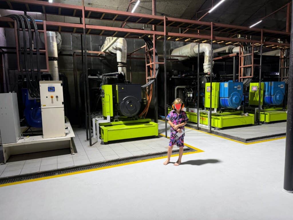 Boy wearing orange ear protection stands barefoot in front of four large generators in a room in Ayada Maldives.