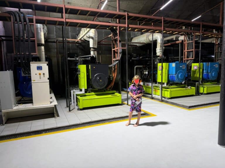 Boy wearing orange ear protection stands barefoot in front of four large generators in a room in Ayada Maldives.