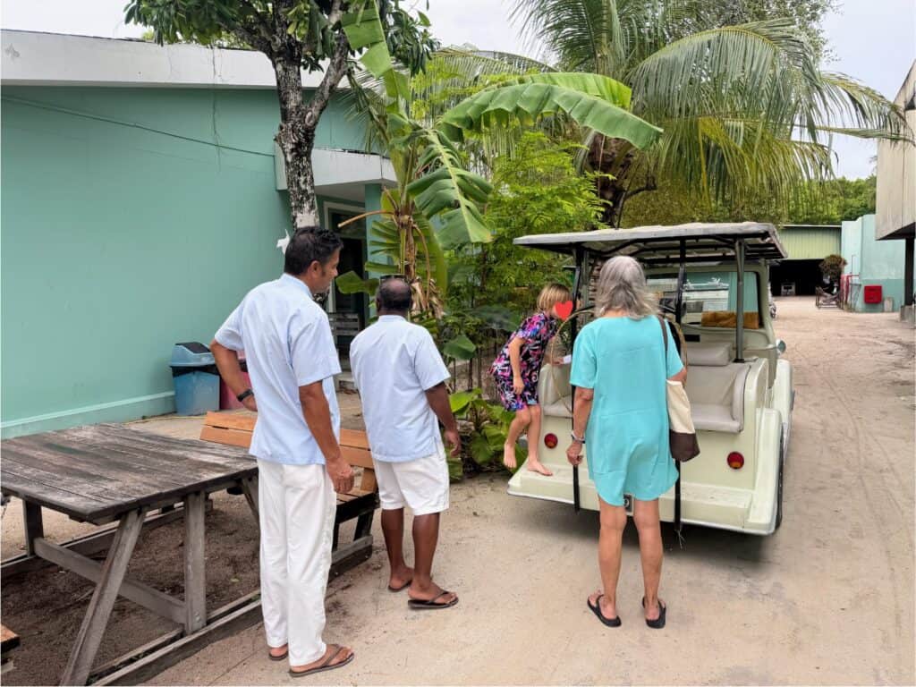 Two staff members, a boy, and a grandmother in a staff area of Ayada Maldives. The boy is getting onto the back of a golf cart while the others look on. Mint colored building on left and other buildings to right and up ahead. Palm trees and other green trees to left of cart and ahead in distance.