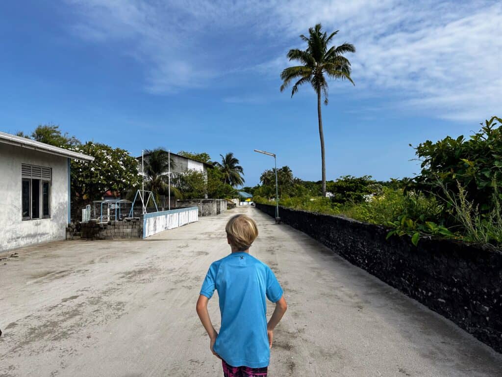 Boy walks down dirt road on Rasgetheemu island in the Maldives. Stone wall borders road on right and a school with a playground is on the left. Tall palm tree ahead on right and other greenery on both sides.