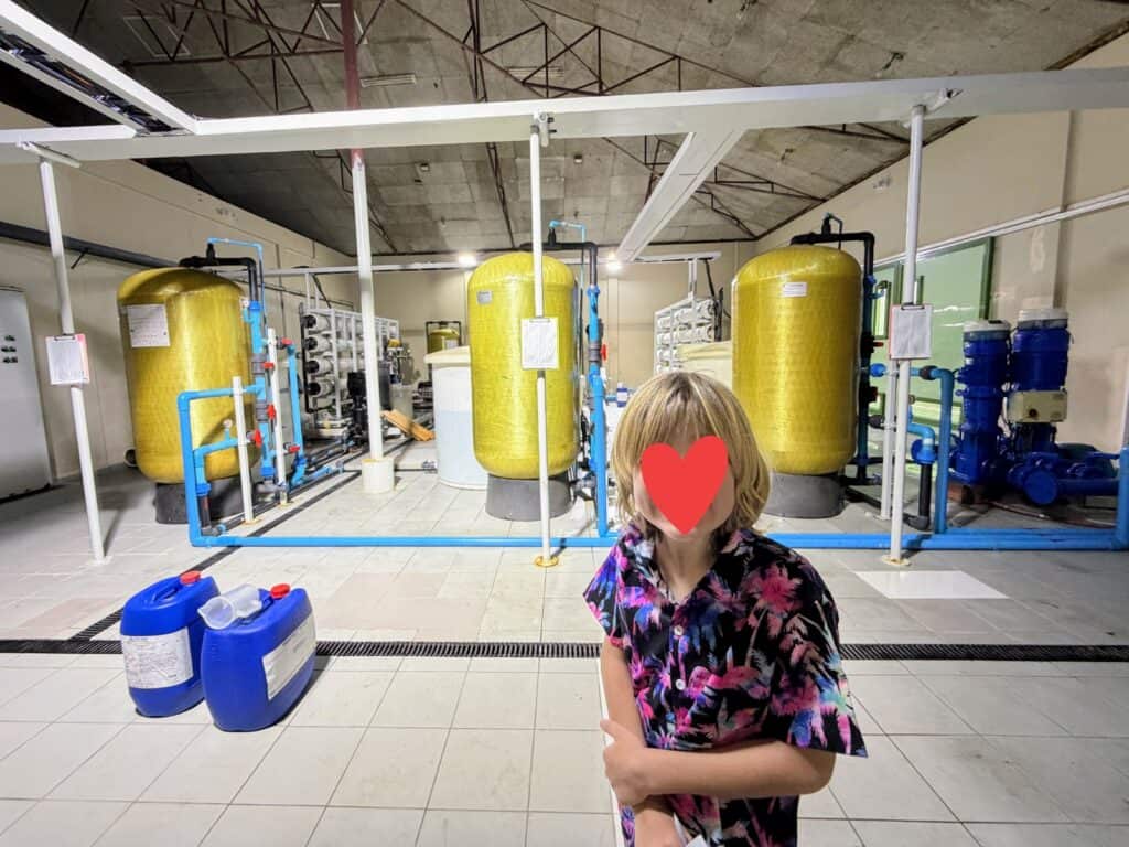 Boy stands in front of water tanks in a water treatment room at Ayada in the Maldives. Two large water jugs on floor on left.