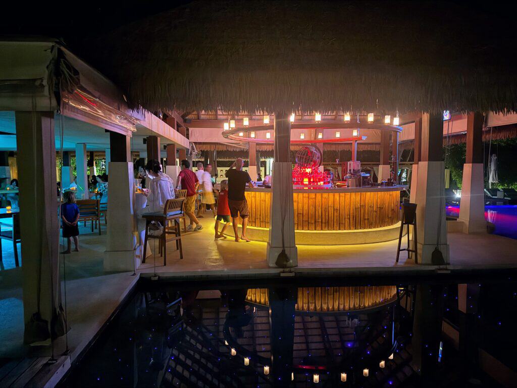 People of all ages are sitting and standing in the bar area at the pool at Ayada Maldives. Thatched roof above the bar. The bottom of the pool has twinkling lights.