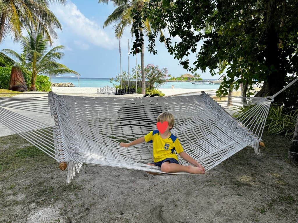 Boy sits in a hammock in a sandy, shaded area at Emerald Faarufushi resort in the Maldives. Outdoor chess set, palm trees, and beach behind him.