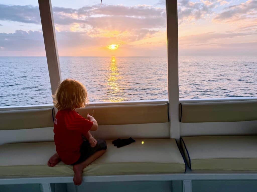 Boy sits on edge of boat and holds fishing line in the ocean in the Maldives at sunset.