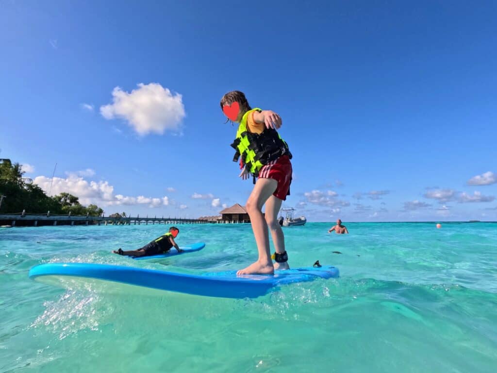 Boy surfing on a foam board in the Maldives. Another child is paddling out and an adult waits in the background. Wooden jetty and overwater villas on left in background.