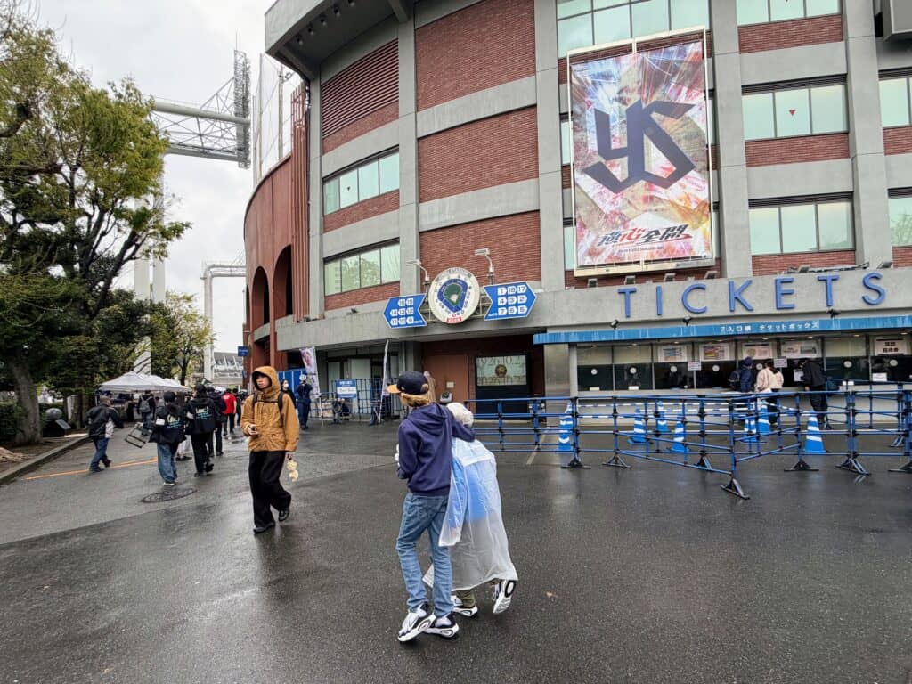Boys walking past ticket booths at Meiji Jingu baseball stadium in Tokyo. It's a cloudy day and there are a few other people around. Large sign with 'YS' on the side of the brick stadium. Trees on left.