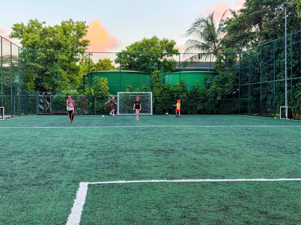 Boys and adults playing soccer on an artificial pitch in the Maldives. Fenced pitch. Green water tanks behind the fence in the background. Trees surrounding the pitch.