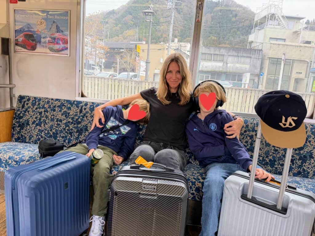 Mother with sons in either arm. They're sitting on a fabric seat in a train. You can see buildings and a hill in the window behind them. They have their small suitcases in front of them.
