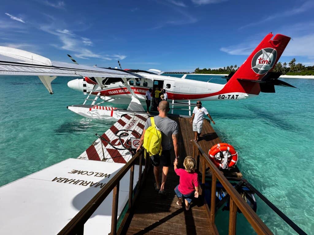 Family walks on wooden jetty toward a seaplane in the Maldives. Island in the background. Turquoise water and a sunny day with whispy clouds.