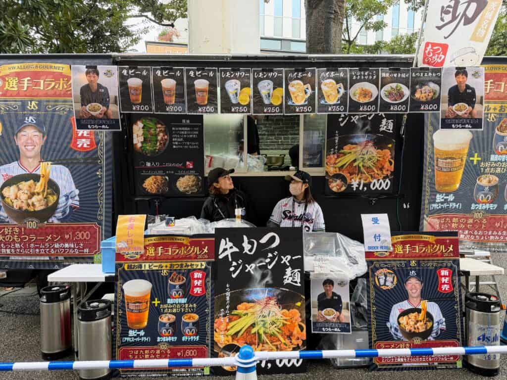 Two women working at an outdoor concessions stand at Meiji Jingu stadium in Tokyo. Beers and Japanese food on the menu.