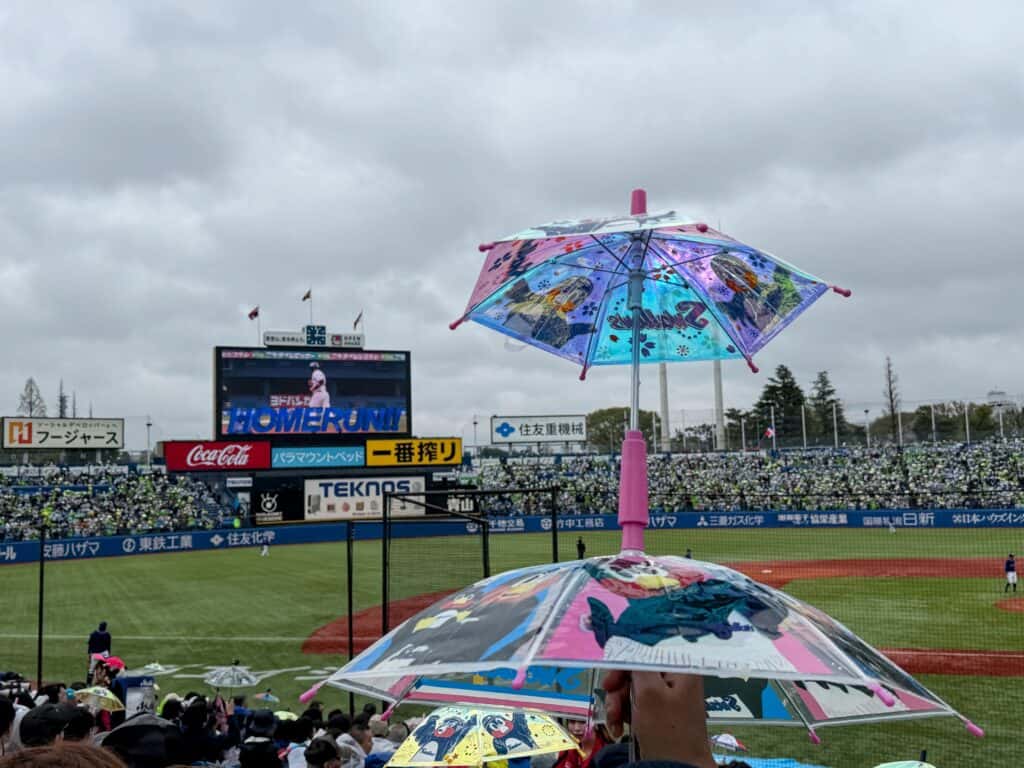 A fan's hand holds up a set of mini umbrellas inside Meiji Jingu baseball stadium on a cloudy day in Tokyo. The scoreboard shows a player running and 'HOMERUN!!'
