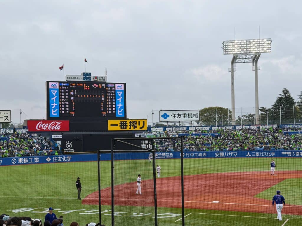 Players on the field at Meiji Jingu baseball stadium in Tokyo, Japan. Digital scoreboard and stadium lights in the background.