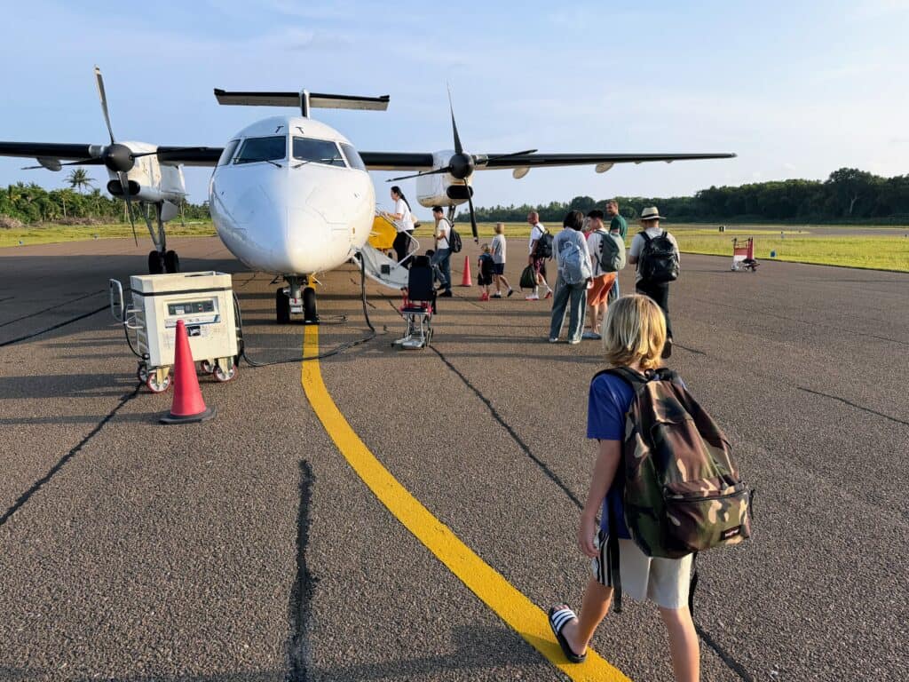 People walking toward and onto a small plan on a runway in the Maldives. Palm trees and forest on either side of the runway.