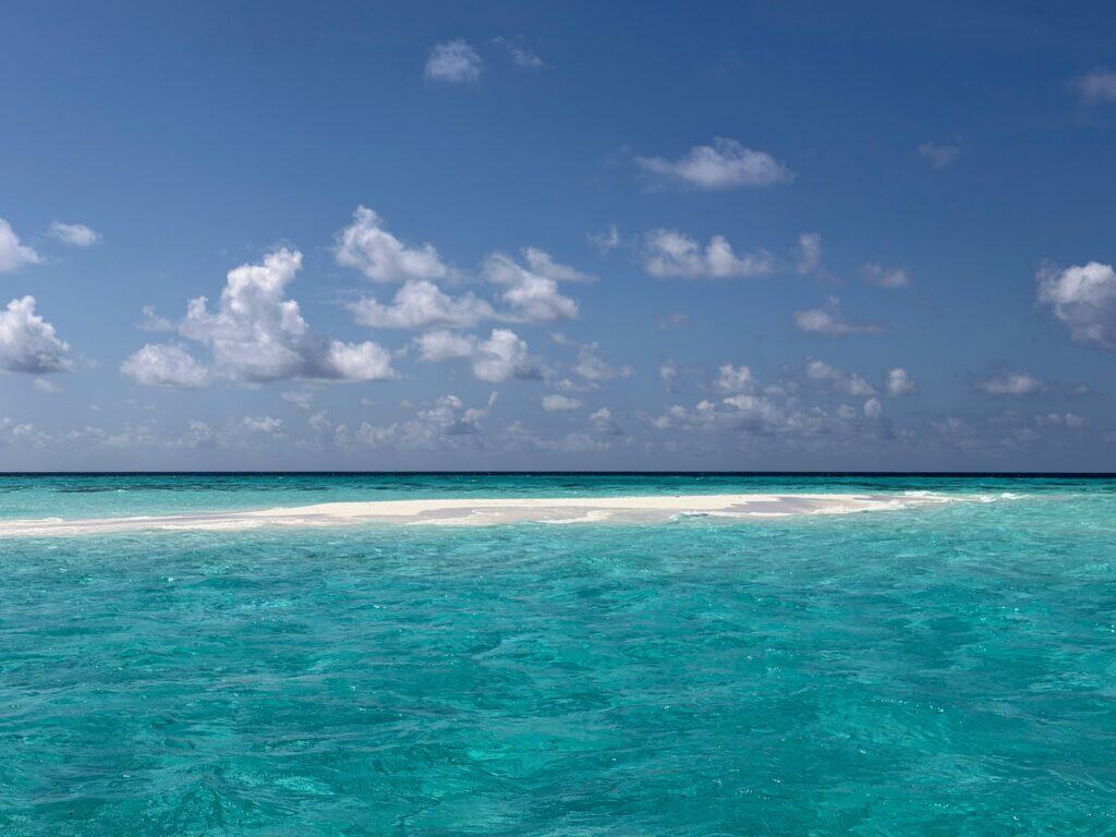 Empty small sandbank surrounded by turquoise water in the Maldives on a sunny day with puffy white clouds.