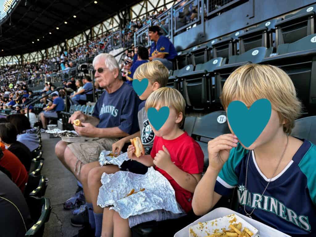 Grandfather and three grandsons sit in stands eating fries and hot dogs at Seattle Mariners baseball stadium.