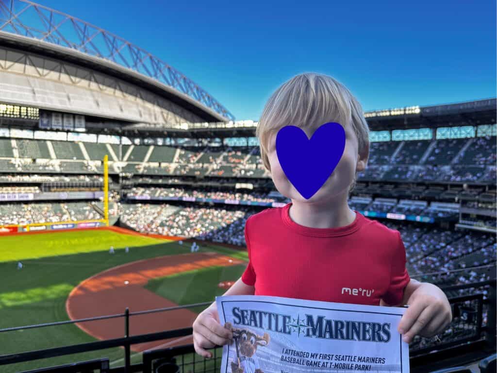 Boy in red shirt holds up certificate for first Seattle Mariners game on a sunny day at Seattle Mariners baseball stadium.