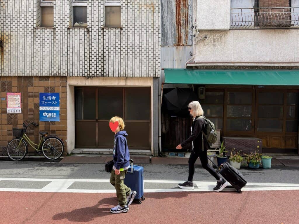 Woman pulls roller suitcase down street in Tokyo. Her grandson walks in front of her and pulls his roller suitcase. Bicycle against wall ahead of them. They are walking to the left.