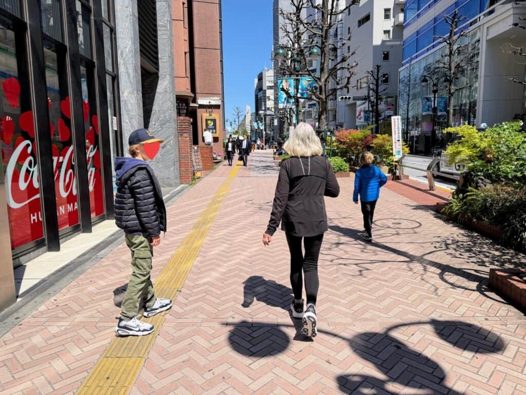 Woman and two grandsons walk down Tokyo pedestrian street on a sunny day. Buildings on either side. They are wearing jackets.
