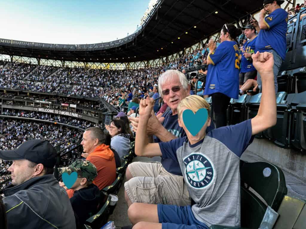 Grandfather claps and grandson raises hands in air to cheer for the Seattle Mariners at T-Mobile Park. The stands are full, and they are sitting in the upper level.
